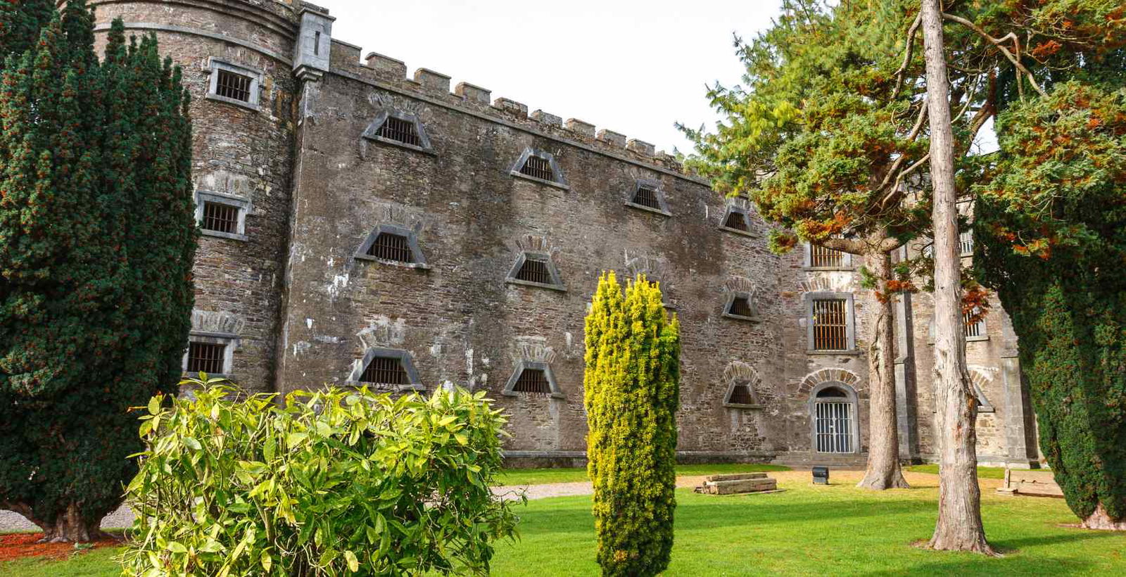 Grassy lawn in front of the historic Old Melbourne Gaol, featuring its stone facade with small windows. The building is surrounded by tall trees, creating a serene and slightly bright atmosphere.