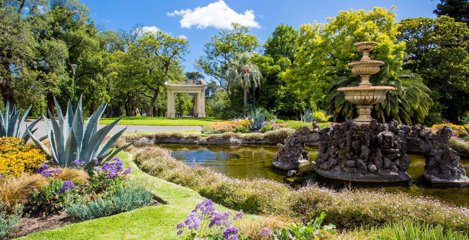 Lush gardens with many plants and lake with fountain surrounded by green grass and a rotunda in the background