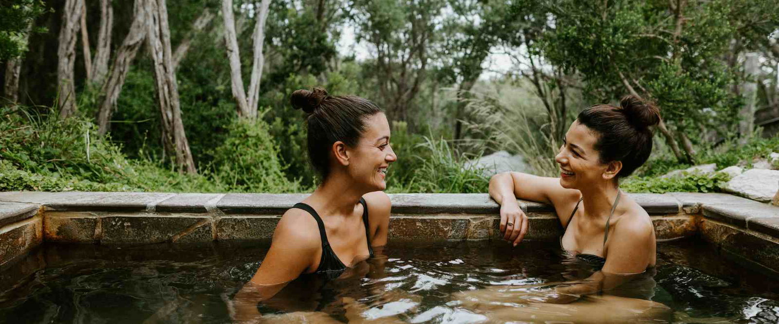 Two women relax in a hot tub surrounded by lush greenery in a serene woodland setting.