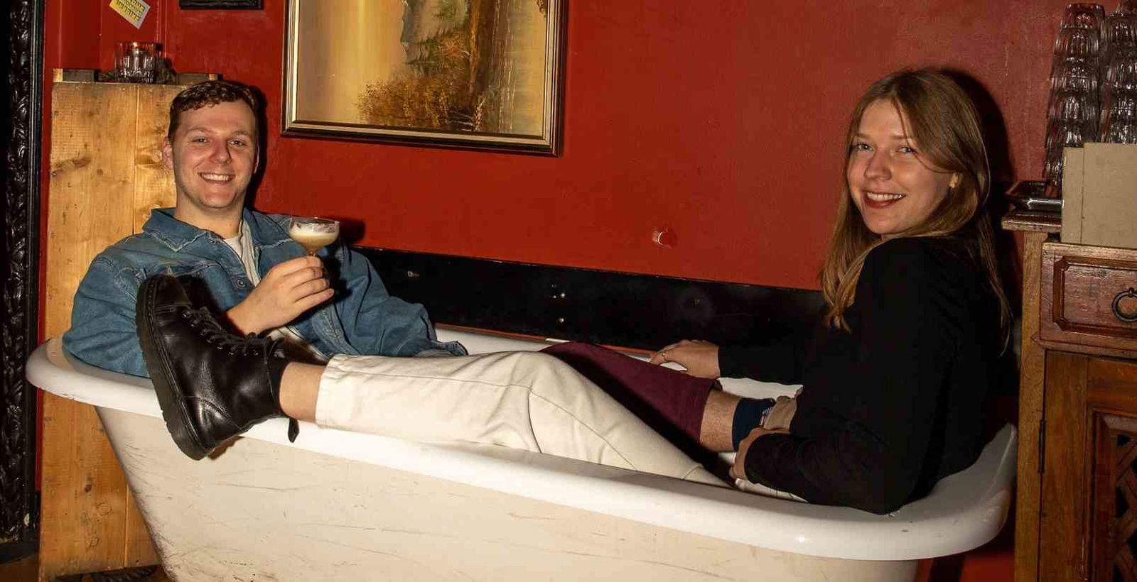 two people sitting in empty prop bathtub in bar while holding cocktails